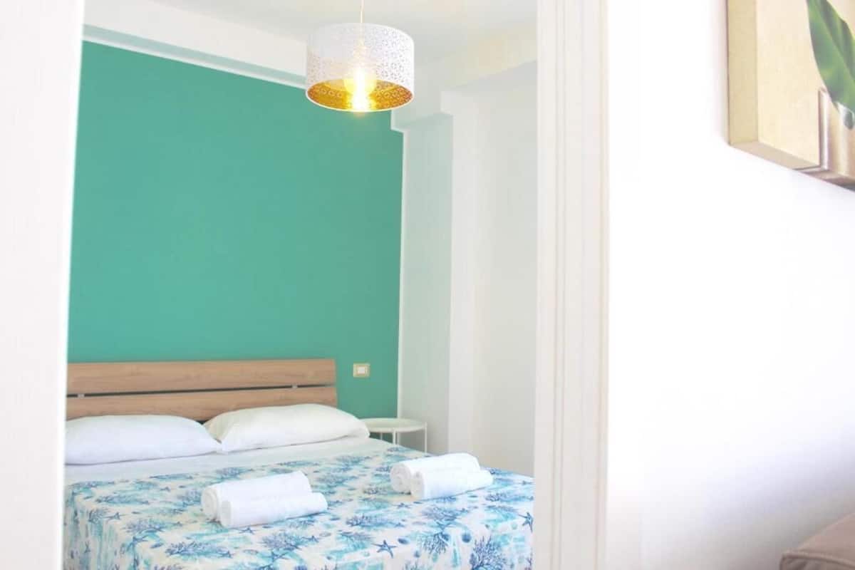 Un dormitorio con pared color turquesa, cabecera de madera, cama con colcha floral, dos toallas enrolladas y una lámpara colgante.