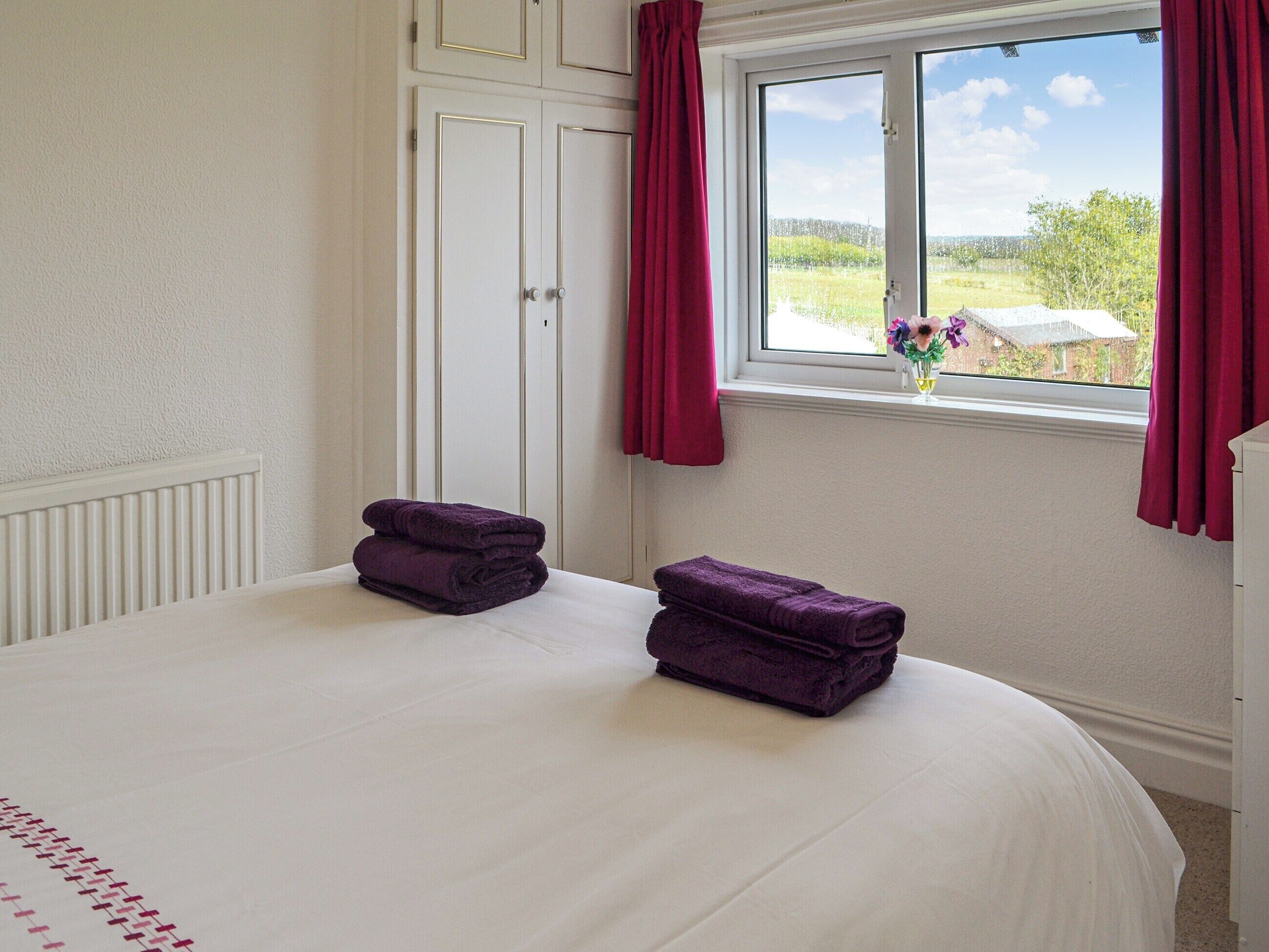 Een slaapkamer met een groot bed, twee opgevouwen paarse handdoeken, een raam met uitzicht op groen en een witte radiator.