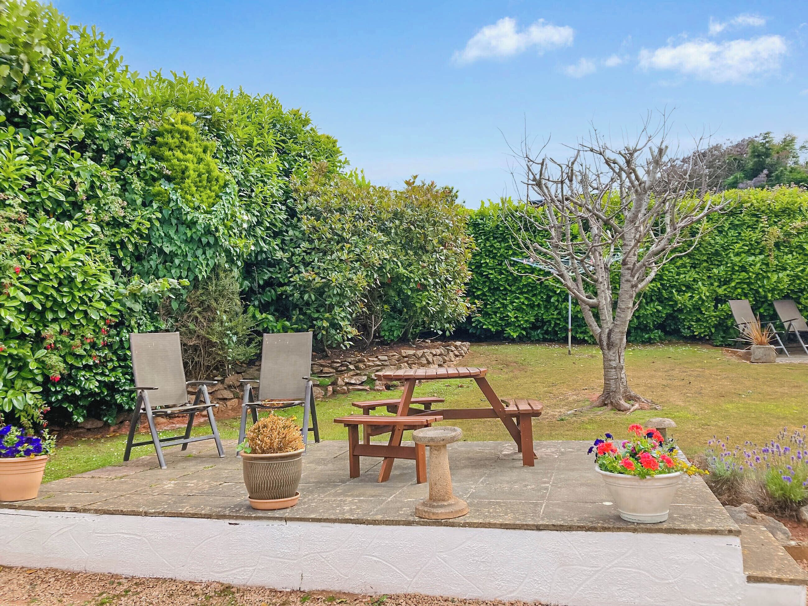 Eine Terrasse mit Tisch und Stühlen, umgeben von üppiger Vegetation und blauem Himmel.