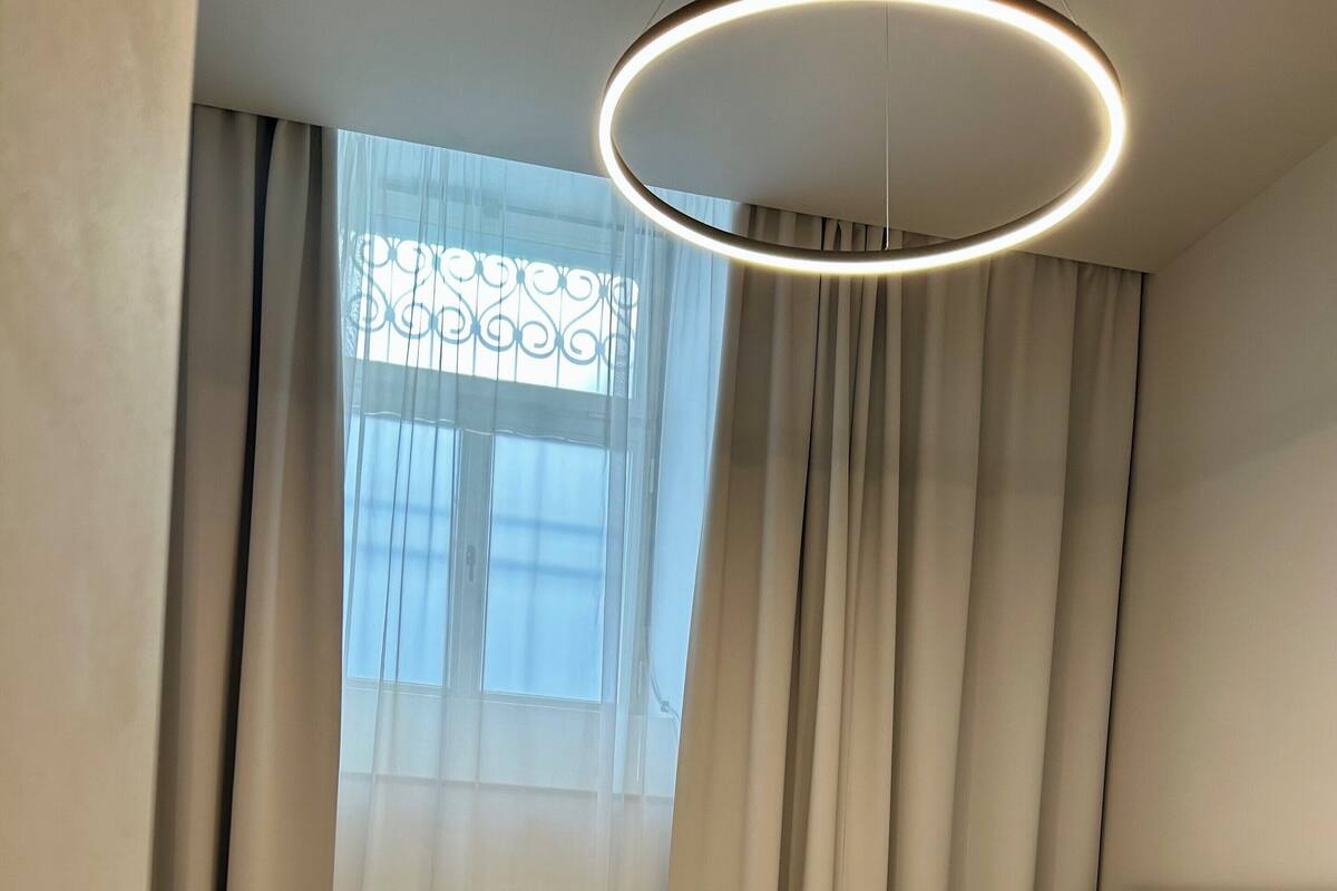 Habitación de hotel con cama, cabecero, ventana con cortinas y lámpara circular.