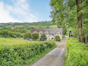 A rural house with a gravel driveway, surrounded by lush greenery and hills in the background.