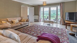 A living room with a sofa, a coffee table, a TV, and a view through large windows. - 4 bedroom accommodation in Llandeilo (Llandeilo)