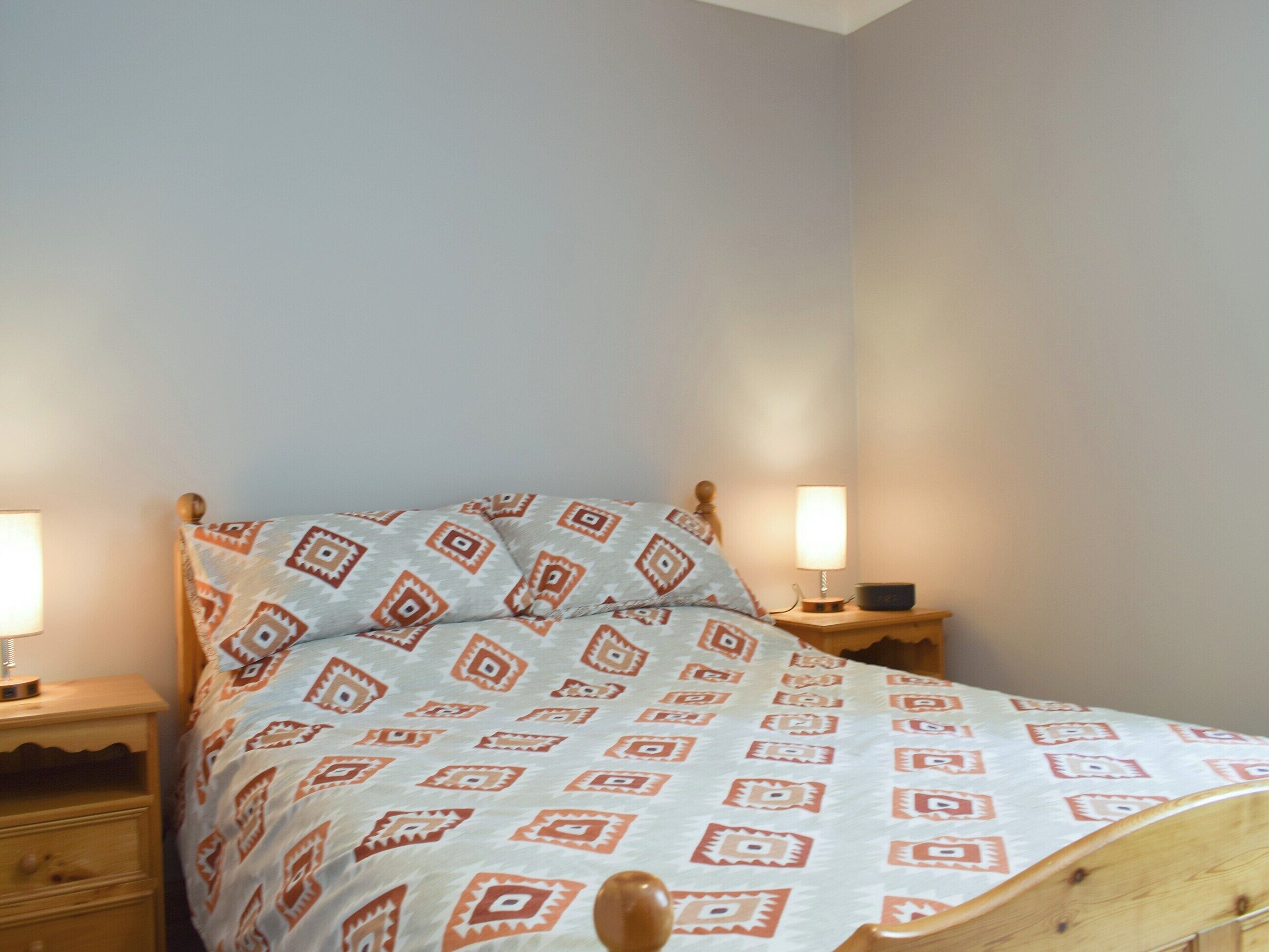 Een bed met een bedsprei met patroon, een houten hoofdbord en twee bedlampen op nachtkastjes van hout.