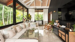 Un salon moderne avec un grand canapé, une table basse et un téléviseur. Il y a des fenêtres avec des rideaux, un ventilateur de plafond et une vue sur la verdure à l’extérieur.