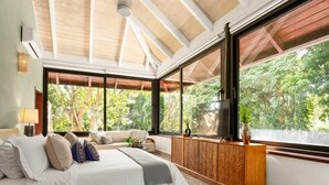 Une chambre avec un grand lit, une armoire en bois et une vue sur les arbres à travers de grandes fenêtres.