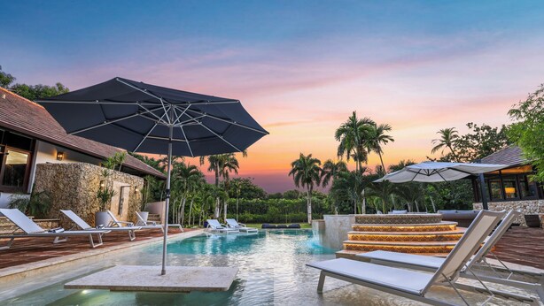 Un espace piscine agrémenté de chaises longues et de parasols, entouré de palmiers et offrant une vue imprenable sur le coucher du soleil.