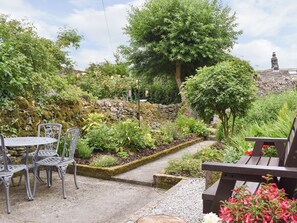 Een tuinpatio met metalen stoelen en een tafel, omringd door weelderige begroeiing en een stenen muur.