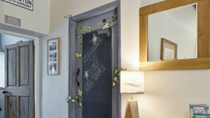 A hallway with a chalkboard door decorated with flowers and a welcome message, a wooden door, a framed picture, a mirror, and a table with a book and a brochure. - Rose Cottage - UK34067 (Grassington)