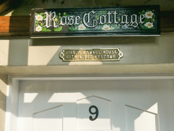 Een huisnummer '9' en een bord erboven met de tekst 'Rose Cottage' en een welkomstdiplomaat.