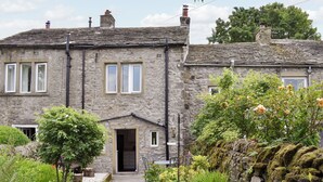 A stone house with a traditional design, a front door, and a small garden with various plants and a moss-covered rock wall. - Rose Cottage - UK34067 (Grassington)