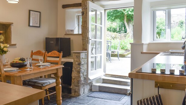 A cozy kitchen with a stone fireplace, a dining area with a wooden table and chairs, and a view of the garden through an open door. - Rose Cottage - UK34067 (Grassington)