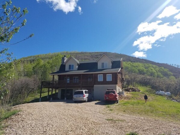 A two-story log cabin with a covered porch, a garage, and a gravel driveway. - Cabin, 4 Bedrooms + Loft, 4.5 Baths, (Sleeps 8-12) Mountain views with trails (Oakley)