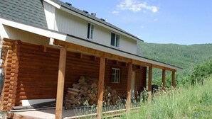 A log cabin with a covered porch, a stack of firewood, and a view of a forested hill. - Cabin, 4 Bedrooms + Loft, 4.5 Baths, (Sleeps 8-12) Mountain views with trails (Oakley)