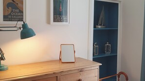 Ein hölzerner Kommode mit Schubladen, ein Spiegel, eine Schreibtischlampe und ein Stuhl. An der Wand befinden sich gerahmte Bilder und ein blaues Regal mit Dekorationen.