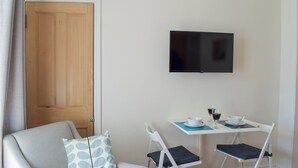 Ein kleines Zimmer mit einer Holztür, einem weißen Sessel mit einem gemusterten Kissen, einem Klapptisch mit zwei Stühlen, einem an der Wand befestigten Flachbildfernseher und einem Tisch mit einer Schüssel, zwei Weingläsern und einer Tasse.