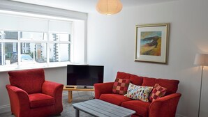 A living room with a red sofa, a red armchair, a grey coffee table, a TV on a wooden stand, a framed painting on the wall, and a floor lamp. - 3 bedroom accommodation in Embleton (Embleton)