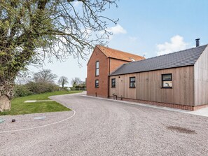 A building with a brick tower and a wooden extension, a gravel driveway, and a grassy area with trees.