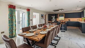 Comedor con mesa de madera dispuesta para seis comensales, rodeada de sillas de cuero, en una cocina moderna con armarios oscuros y piso de mármol.