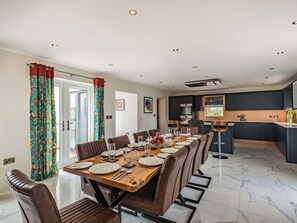A dining area with a wooden table set for six, surrounded by leather chairs, in a modern kitchen with dark cabinetry and a marble floor.