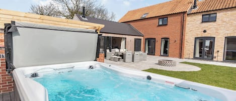 A hot tub in a backyard with a patio area, a brick house, and a covered seating area.