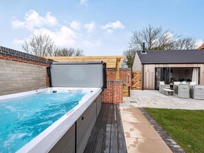 Een hottub naast een houten terras, een bakstenen muur en een overdekte patio met tuinmeubilair.