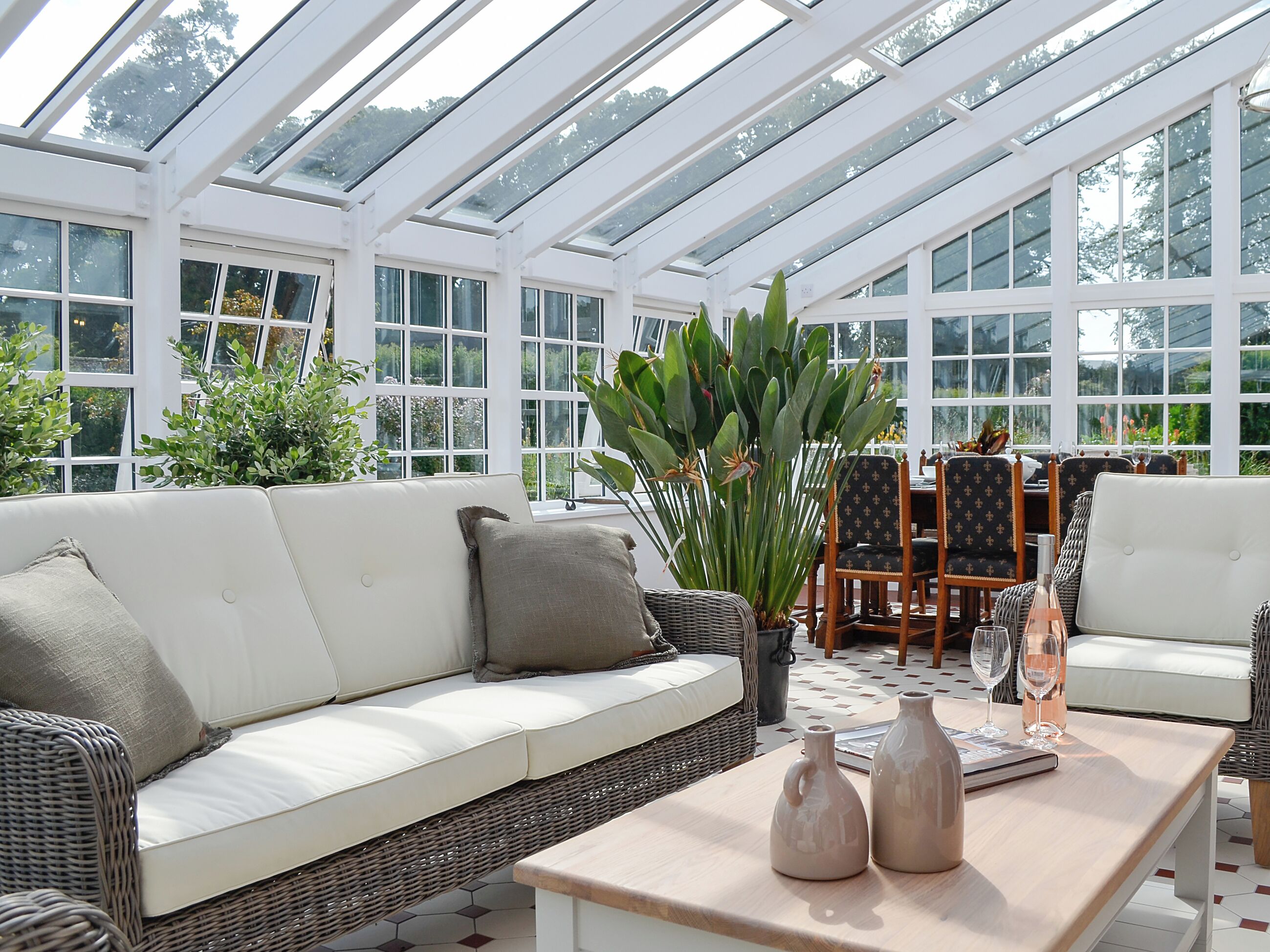 Ein Wintergarten mit einem weißen, gepolsterten Sofa, Korbstühlen, einem Tisch mit Vasen und einer großen Topfpflanze.
