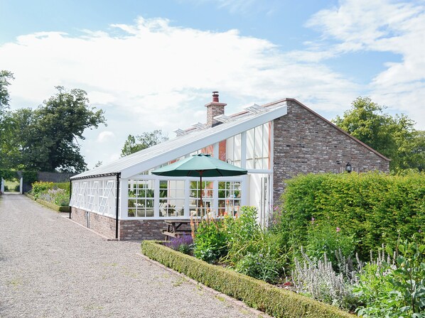 A stone building with a conservatory, a gravel driveway, and lush garden vegetation.