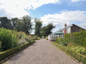 A gravel pathway leading to a traditional house with a green roof, surrounded by lush greenery and trees.