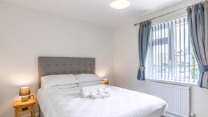 Een net opgemaakt bed met witte lakens, een houten nachtkastje met een lamp, en een raam met blauwe gordijnen.