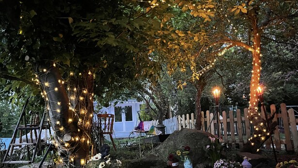 Un jardin agrémenté de jeux de lumière décoratifs, une clôture en bois et un bâtiment blanc en arrière-plan.