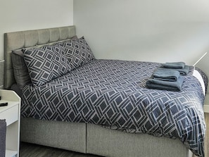 Ein ordentlich bezogenes Bett mit einem geometrisch gemusterten Überwurf und Kissen, ein Nachttischchen mit einer Lampe und ein gefaltetes Handtuch auf dem Bett.