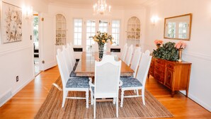 Un comedor con una gran mesa de madera, sillas blancas, una araña y un jarrón con flores.