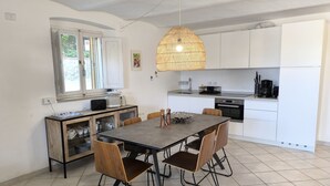 Une cuisine moderne avec des meubles blancs, une table à manger entourée de quatre chaises, un luminaire suspendu et une fenêtre donnant sur un paysage verdoyant.