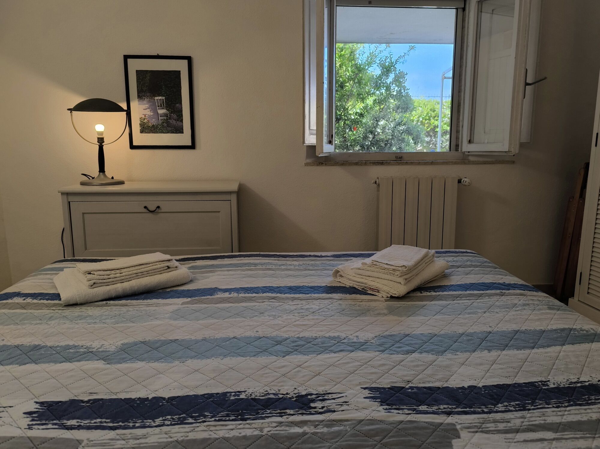 Un dormitorio con cama, lámpara de noche, un cuadro enmarco y ventana con vista a follaje.
