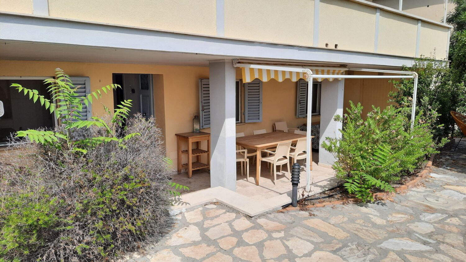 Un espace extérieur abrité, aménagé avec une table et des chaises, entouré de plantes et doté d’un patio dallé de pierres.