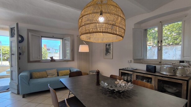 Une cuisine moderne avec une table à manger, des chaises, un grand luminaire en fibres tressées et une vue sur l’extérieur grâce à de grandes fenêtres.