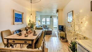 A cozy living room with a wooden dining table, chairs, a sofa, and a television. There is a window with a view of the sea, and a plant on the table. - 2 bedroom accommodation in Ilfracombe (Ilfracombe)
