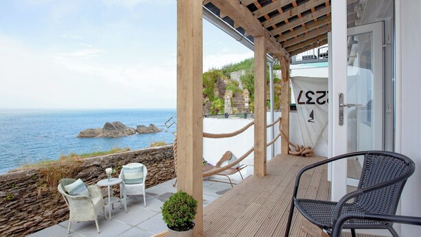 A covered outdoor seating area with a view of the sea, featuring a wooden pergola, white wicker chairs, and a black metal chair. - 2 bedroom accommodation in Ilfracombe (Ilfracombe)