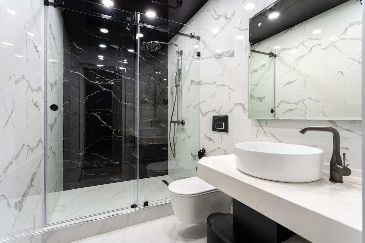 Un baño moderno con ducha envuelta en vidrio, un lavamanos blanco y pared con azulejos de mármol.