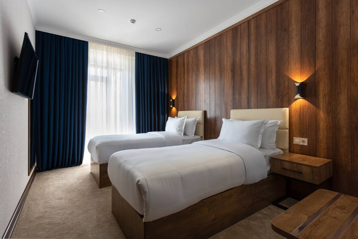 Una habitación de hotel con dos camas, paneles de madera, un televisor montado en la pared y una mesita de noche con lámpara.