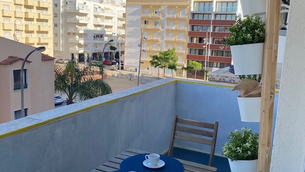 Un balcón con mesa y sillas, plantas en macetas y vistas a edificios al fondo.