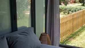 A bedroom with a large window, a sofa, a small table, and a view of a wooden fence and greenery outside.