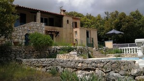 Una casa de piedra de dos pisos con una escalera de piedra que conduce a la entrada, rodeada de exuberante vegetación y una zona de piscina.