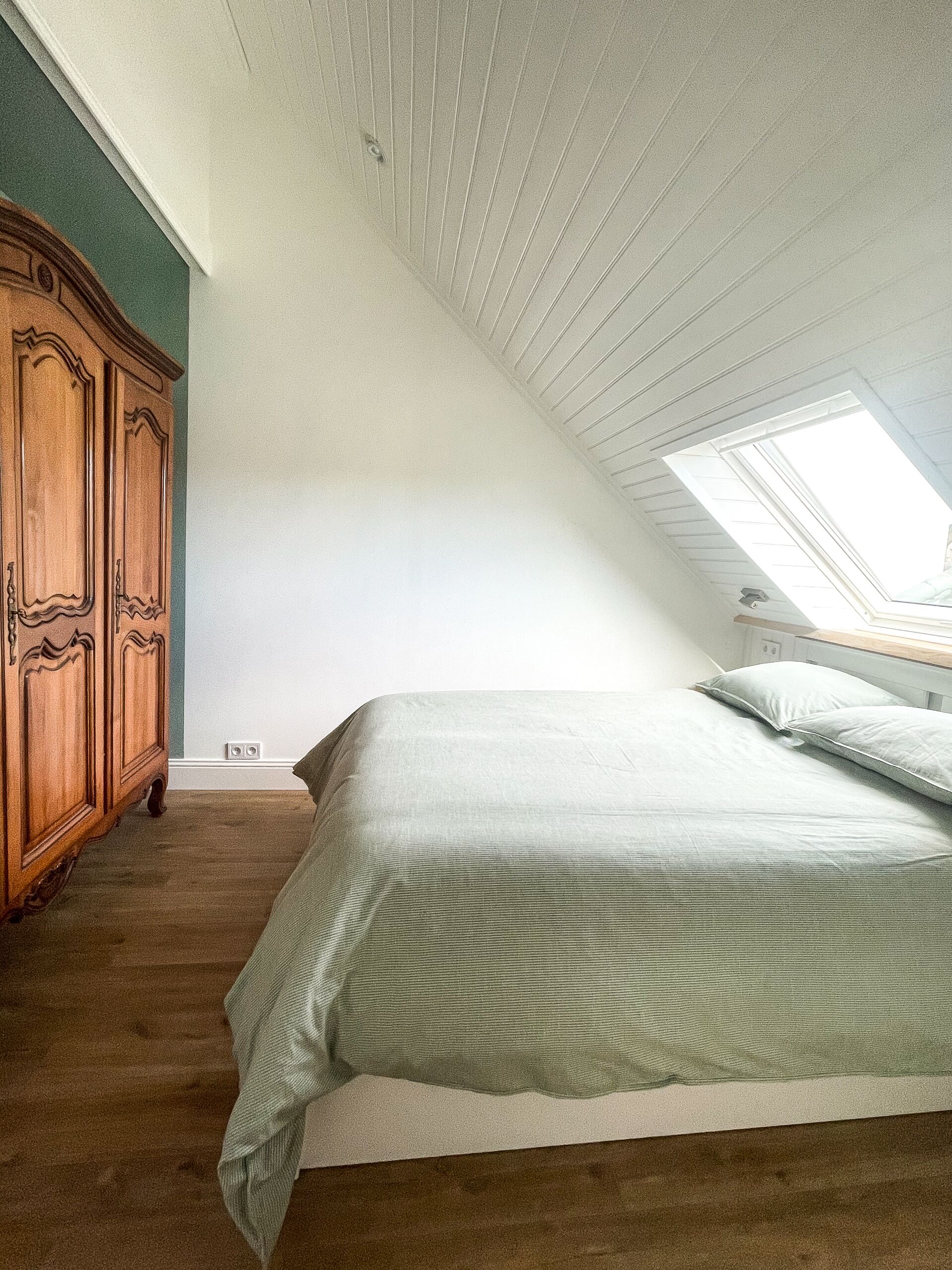 Een slaapkamer met een schuine kap, een dakraam, een houten garderobe en een bed met kussens.