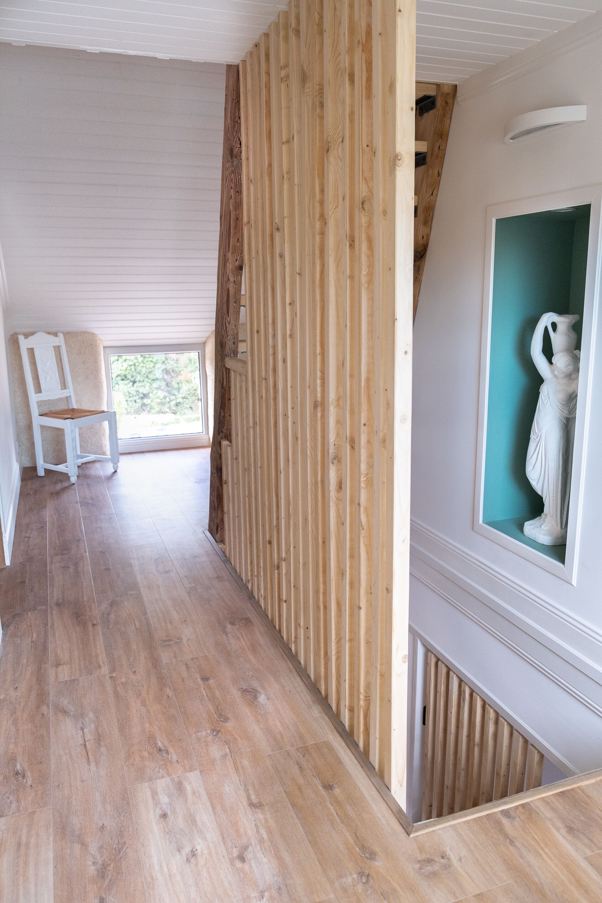 Een kamer met houten vloer, een witte stoel, een houten scheidingswand en een lijst met een kunstwerk aan de muur.