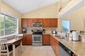 Une cuisine moderne avec des meubles en bois, des appareils en acier inoxydable, un plan de travail en granit et un coin repas comprenant une table et des chaises blanches.
