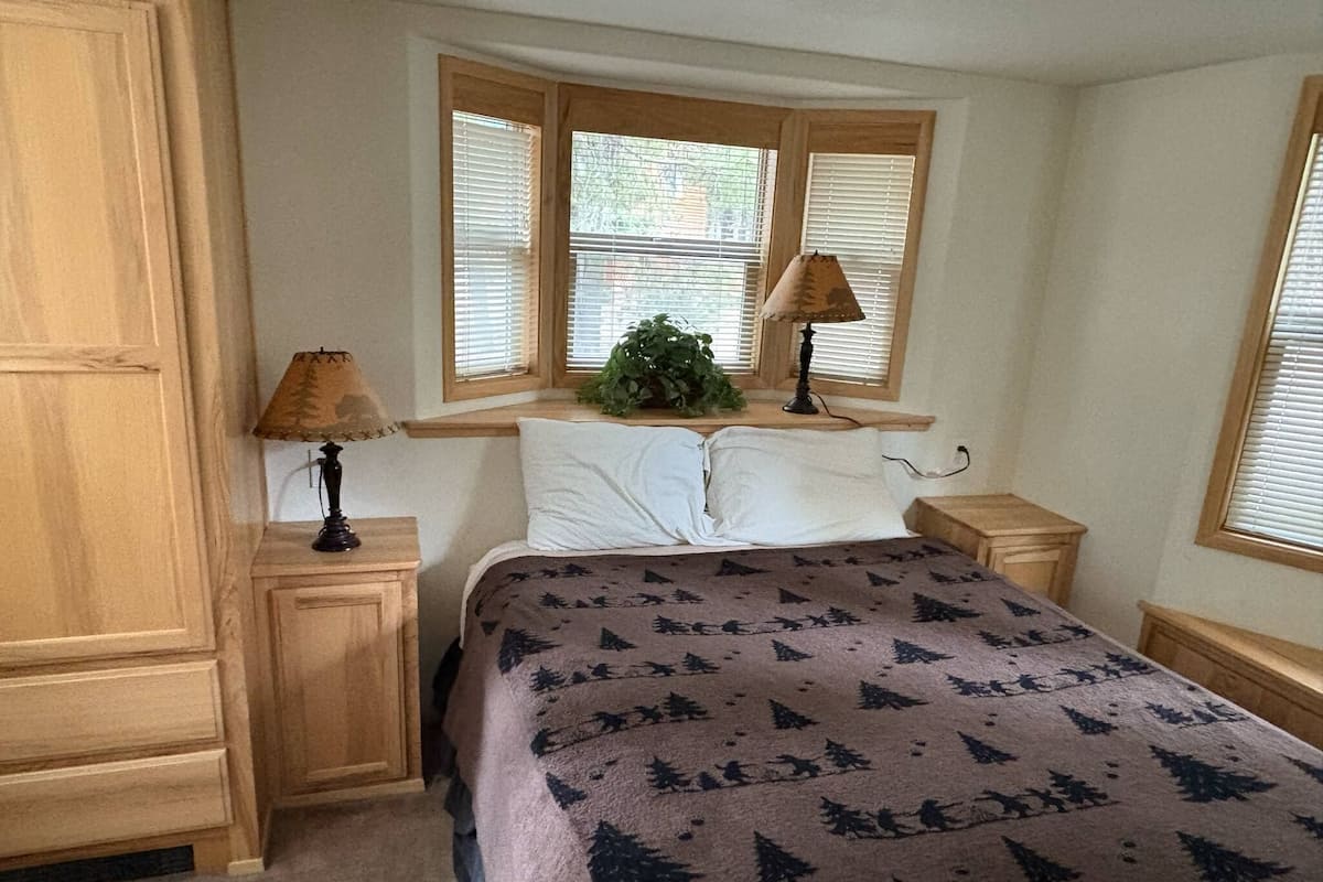 A bedroom with a bed, two bedside tables, a window with blinds, a lamp on each table, and a plant on the windowsill.