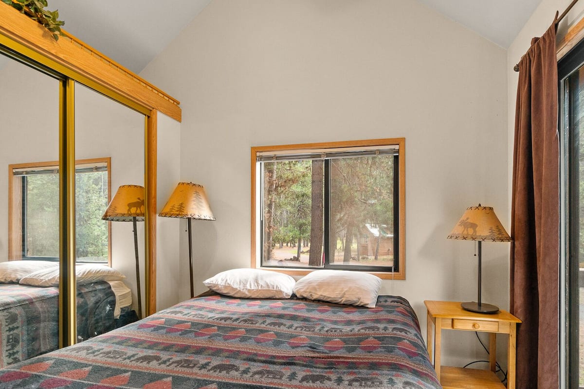 A bedroom with two beds, a large mirror, a window with a view of trees, and a lamp on a bedside table.