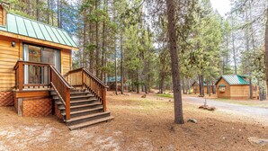 Una cabaña con fachada de madera, una escalera que sube a una terraza, y un bosque de fondo.