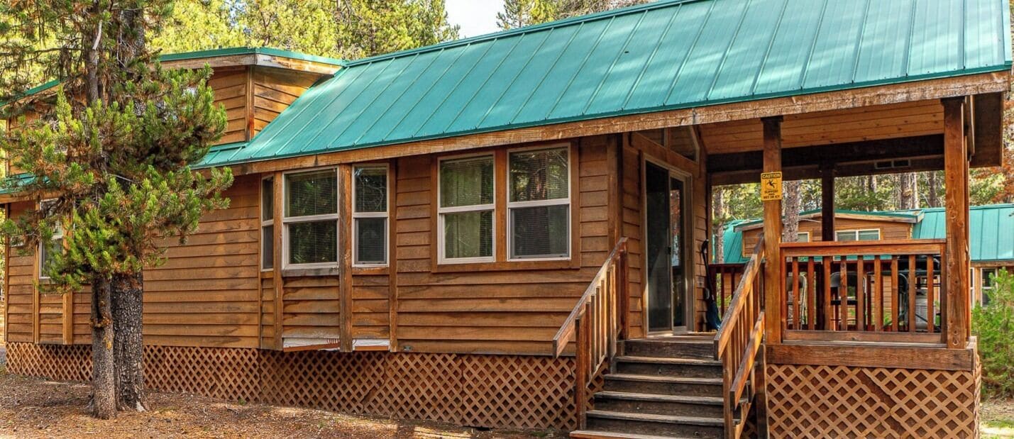 Une cabane en bois avec un toit en métal vert, une véranda dotée d’une balustrade en bois et un escalier menant à l’entrée.